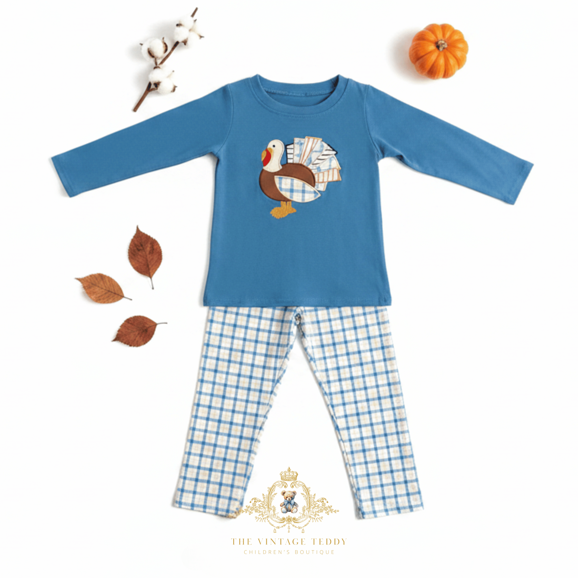 A toddler boys two-piece matching set featuring a blue long-sleeve top with a turkey embroidery design and matching blue-and-white plaid pants, styled flat-lay on a white background with autumn props including: mini pumpkin, cotton stem, brown leaves and The Vintage Teddy's logo watermark at the bottom.