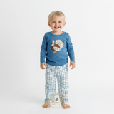 A smiling toddler boy standing barefoot against a plain white studio background, wearing a blue long-sleeve top with an embroidered turkey and blue-and-white checkered long pants.