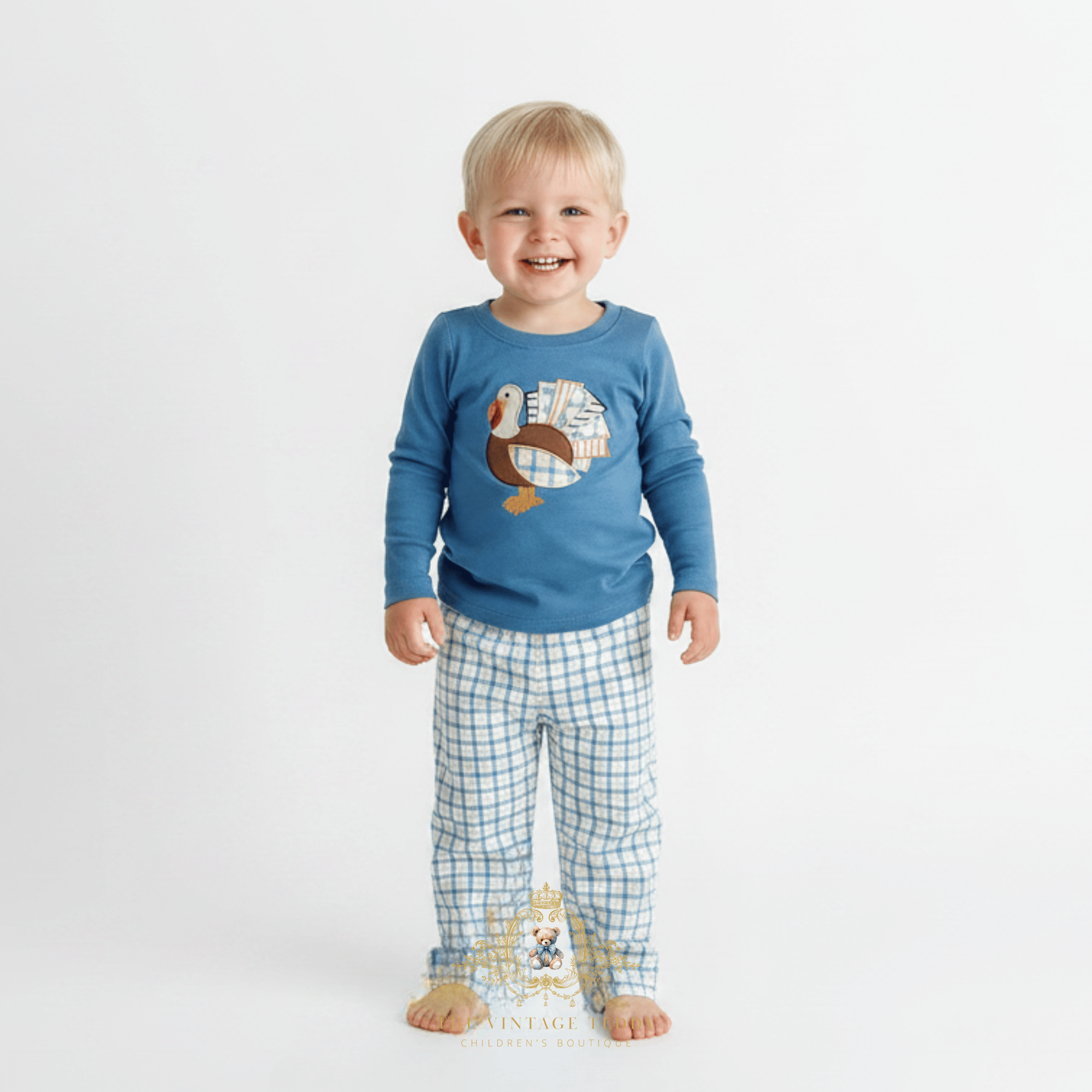 A smiling toddler boy standing barefoot against a plain white studio background, wearing a blue long-sleeve top with an embroidered turkey and blue-and-white checkered long pants.
