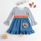 A toddler girl's long-sleeve dress with a navy and white striped bodice, a coral orange waistband, and a blue skirt featuring a turkey embroidered applique. The dress is styled in a flat lay with cotton bolls, autumn leaves, and a small pumpkin on a white background.
