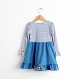 The back of a children's long-sleeve dress hanging on a wooden hanger against a plain white wall. The top half has a navy and white horizontal stripe pattern, while the bottom half is a solid medium blue with a ruffled hem.