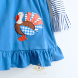 A close-up of a blue children's dress with a ruffled hem and a striped sleeve, featuring a turkey applique patch on the front. The dress is shown against a white background with a The Vintage Teddy's watermark at the bottom.