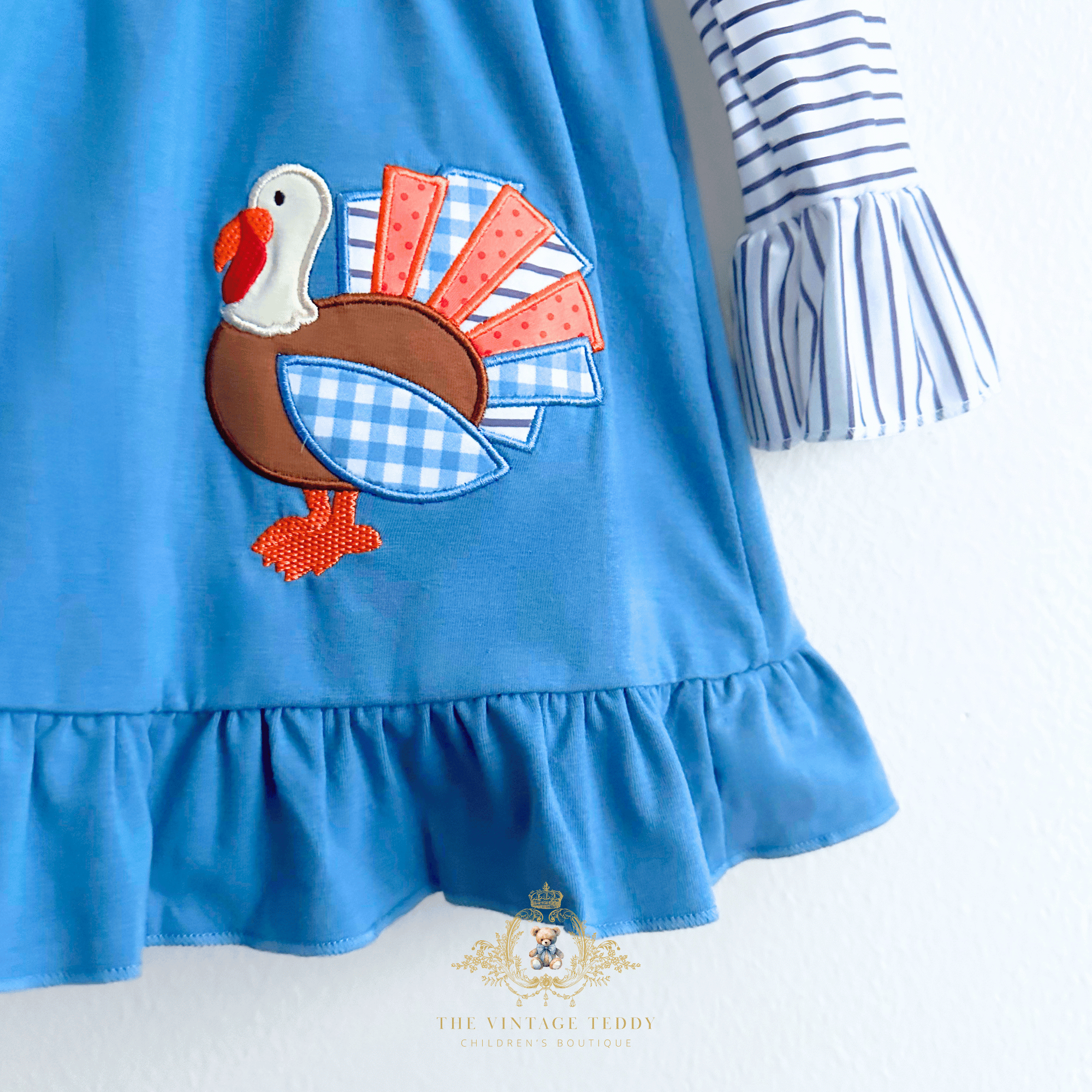 A close-up of a blue children's dress with a ruffled hem and a striped sleeve, featuring a turkey applique patch on the front. The dress is shown against a white background with a The Vintage Teddy's watermark at the bottom.