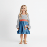 A young girl with long golden hair stands barefoot against a plain white studio background wearing a long-sleeve dress with navy blue and white horizontal stripes on the top and sleeves, a coral-red polkadot waistband, and a cornflower blue skirt featuring a turkey applique.
