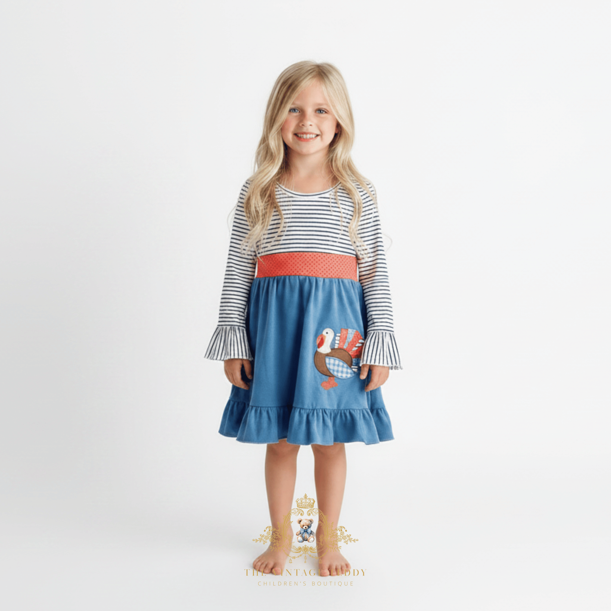 A young girl with long golden hair stands barefoot against a plain white studio background wearing a long-sleeve dress with navy blue and white horizontal stripes on the top and sleeves, a coral-red polkadot waistband, and a cornflower blue skirt featuring a turkey applique.