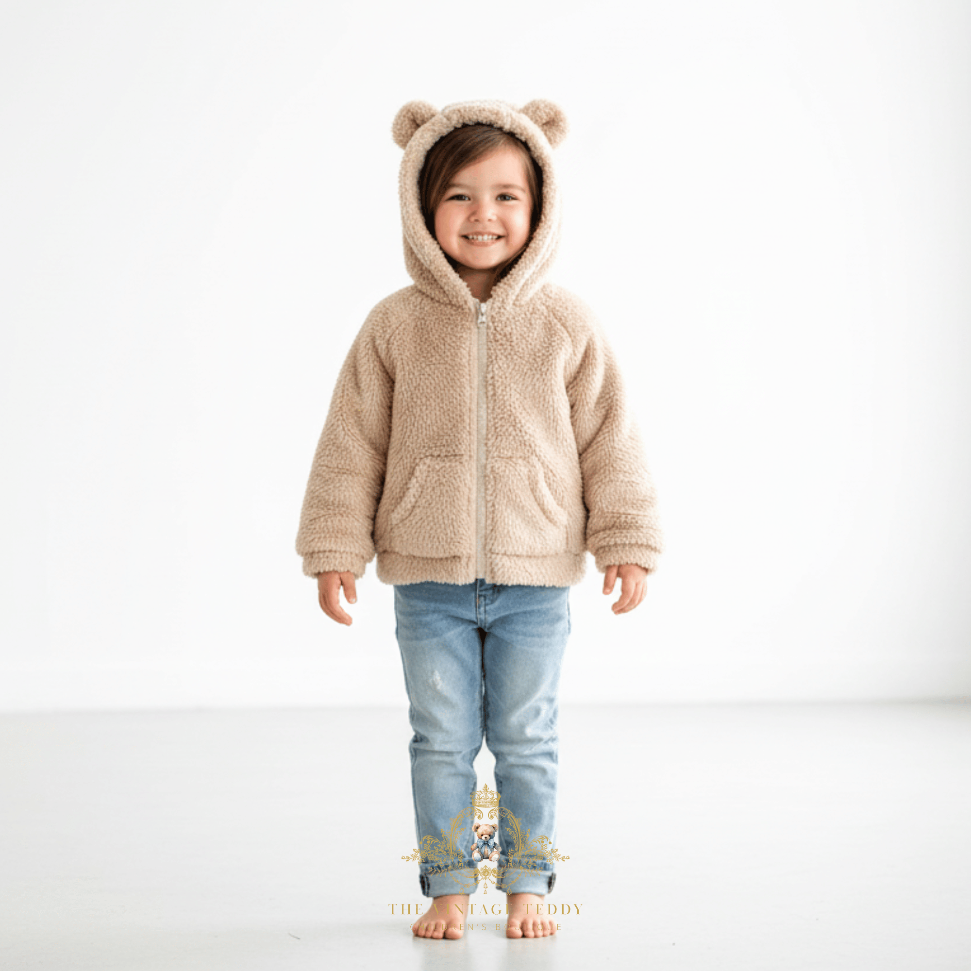 A young child, happy little girl wearing a beige sherpa fleece zip-up hooded jacket with bear ears on the hood, paired with light blue jeans, standing against a plain white studio background.