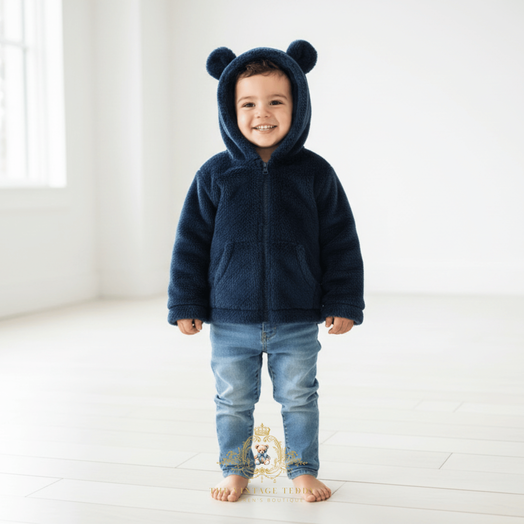 A smiling toddler boy stands barefoot in a bright white room wearing a comfy navy blue fleece sherpa zip-up hooded jacket with bear ears on the hood and light blue jeans.