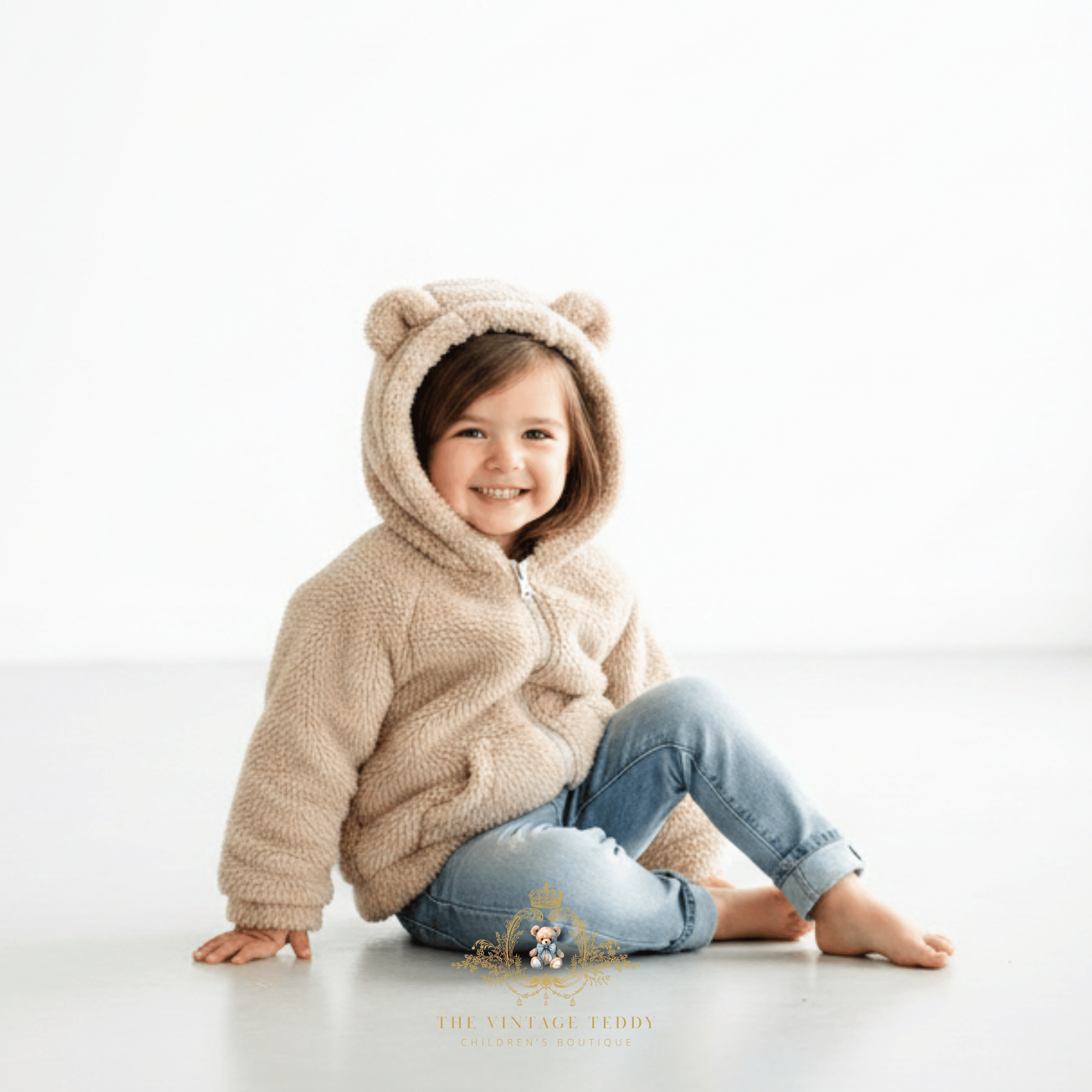 A young child, little girl with brown hair tucked into a hoodie, smiling and sitting on a white floor wearing a beige cream color sherpa fleece hoodie jacket with bear ears on the hood and light blue jeans.