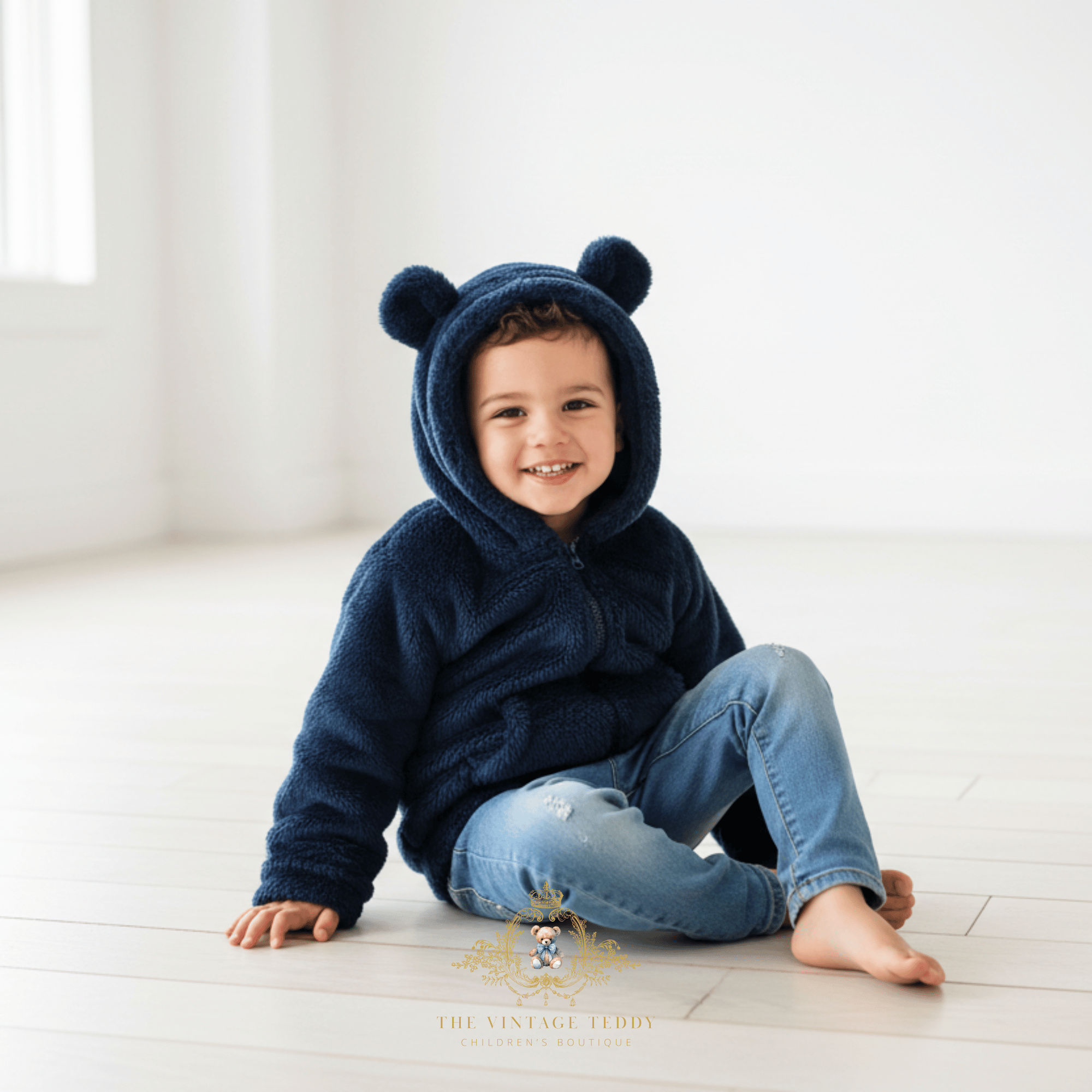 A smiling young child sits barefoot on a light wood floor wearing a plush navy hooded coat with bear ears and blue jeans. Sitting in a bright, minimal room with soft natural light.