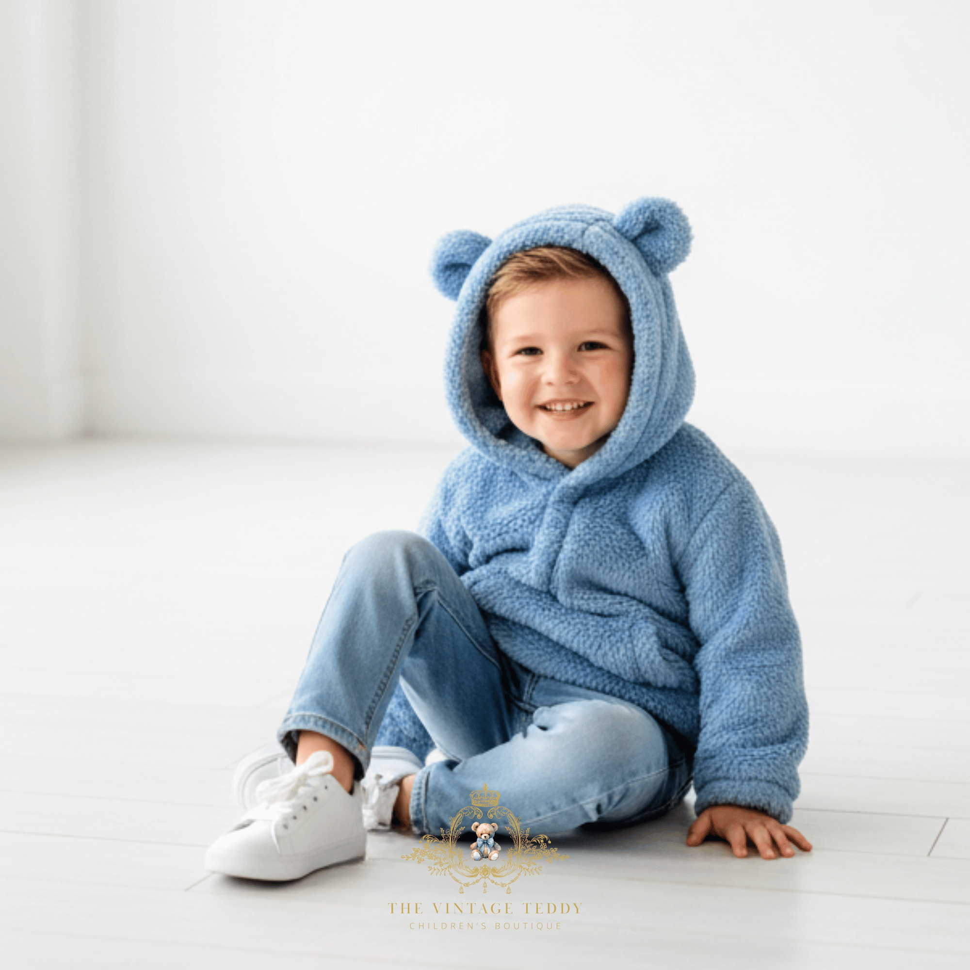 A smiling toddler boy sits on a white floor wearing a plush blue hooded outfit with bear ears and matching blue jeans and white sneakers. A watermark at the bottom reads The Vintage Teddy.