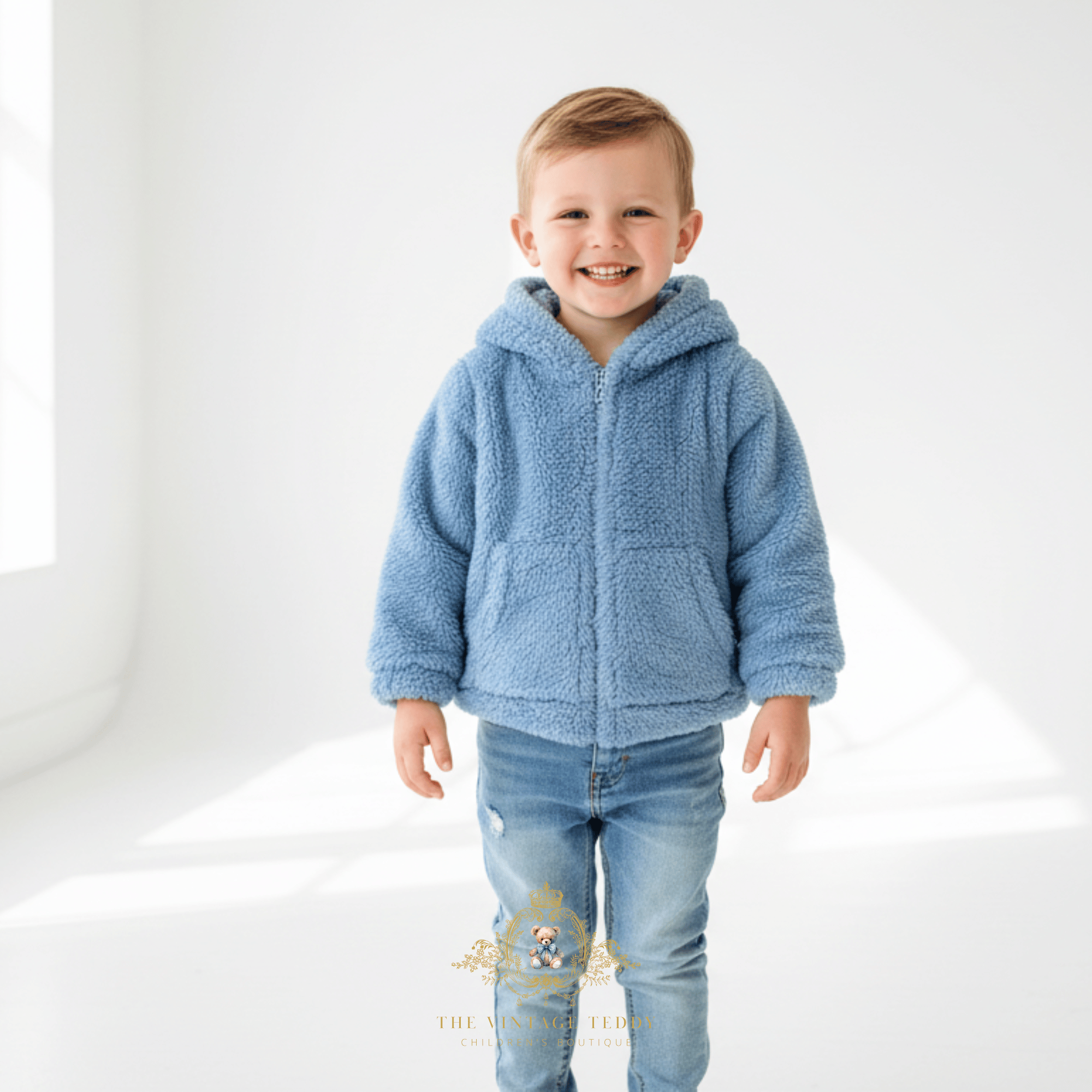 A young child smiling and wearing a sky blue sherpa zip-up hooded jacket with front pockets, paired with blue jeans, standing in a bright white studio setting.