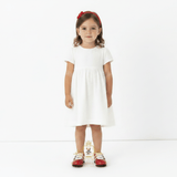 A young girl stands against a plain white studio background wearing a short-sleeve white dress, a red headband, and red-and-white kids sneakers.