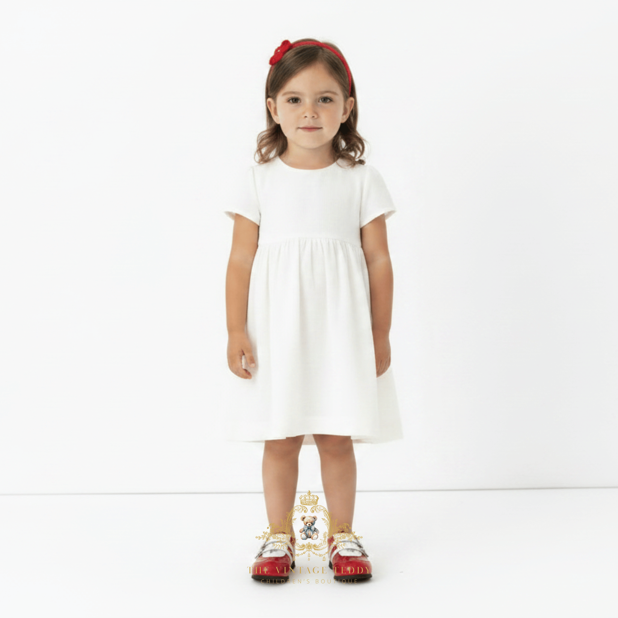 A young girl stands against a plain white studio background wearing a short-sleeve white dress, a red headband, and red-and-white kids sneakers.