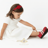 A young girl in a white dress sits on a white floor, pointing at a red children' shoes with white stripes. She is wearing a red flower headband.