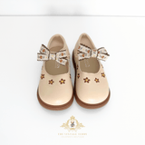 A pair of beige Mary Jane style shoes for young girls, featuring embroidered floral details and matching fabric bows, shown against a plain white background with a brand watermark at the bottom.