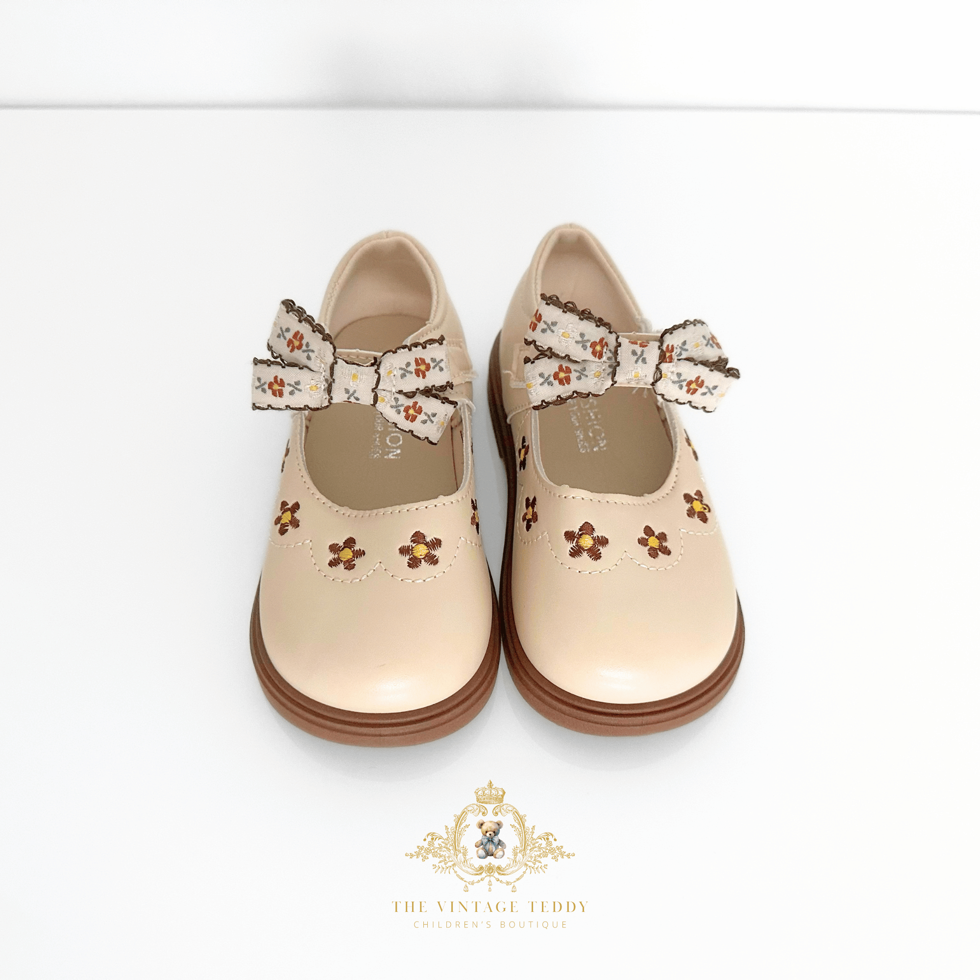 A pair of beige Mary Jane style shoes for young girls, featuring embroidered floral details and matching fabric bows, shown against a plain white background with a brand watermark at the bottom.
