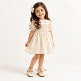 A young girl wearing a cream dress with a bow in her brown wavy hair and Mary Jane flat shoes for kids on a white studio background with The Vintage Teddy's brand logo bottom center.