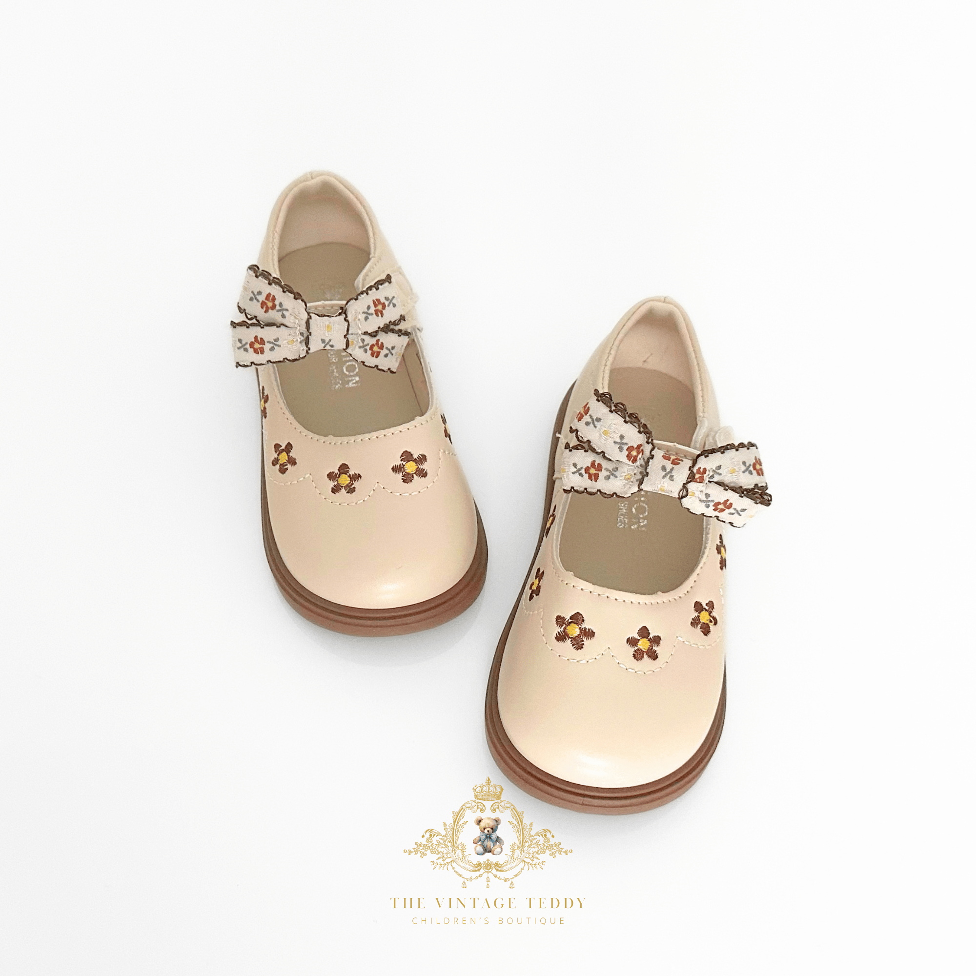 A pair of beige Mary Jane style children's shoes with embroidered floral details and bow accents, displayed on a clean white background with a The Vintage Teddy brand logo at the bottom.