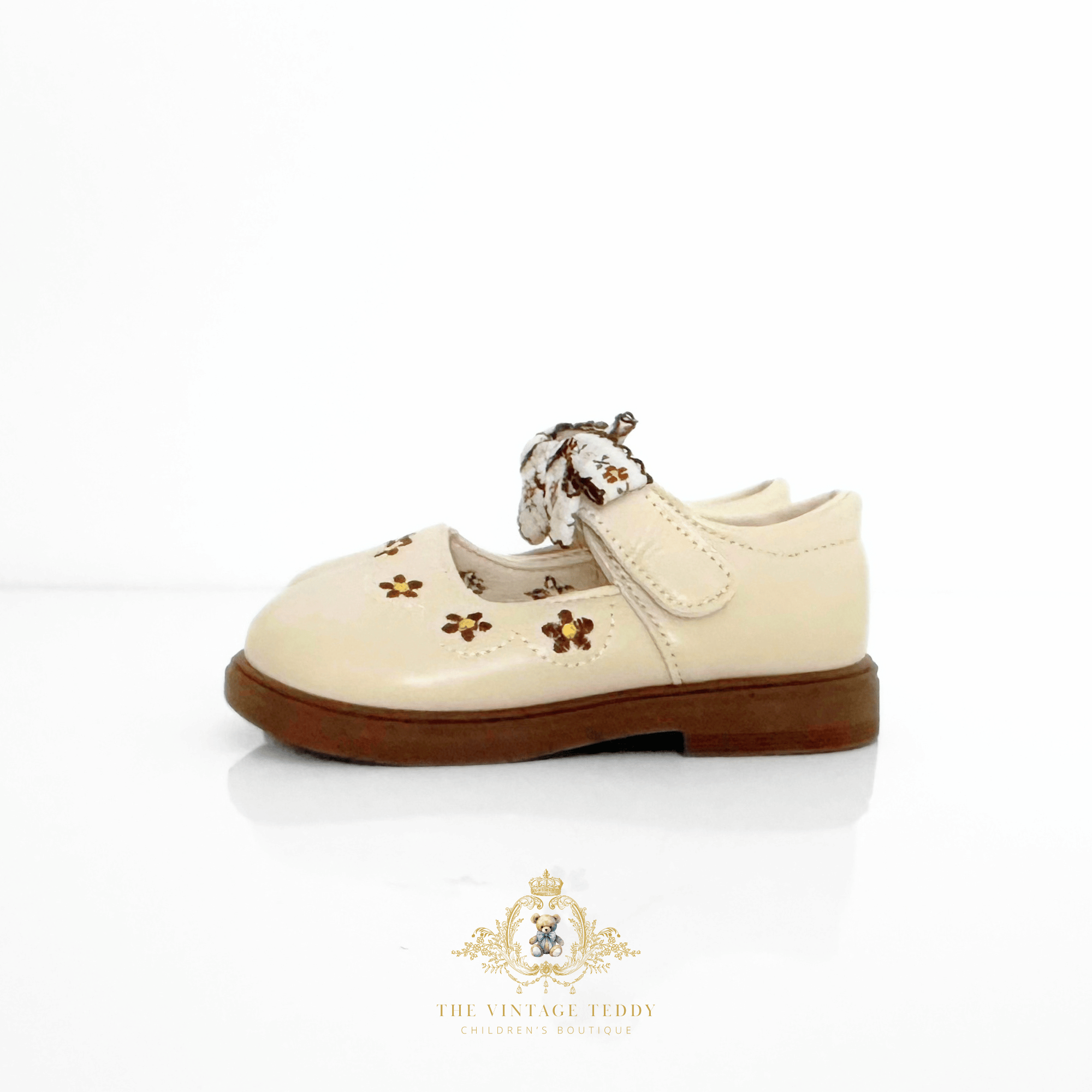 A cream-colored leather toddler girl's shoe for kids with brown sole, decorated with embroidered floral details and a matching floral bow on the strap, shown against a white background.