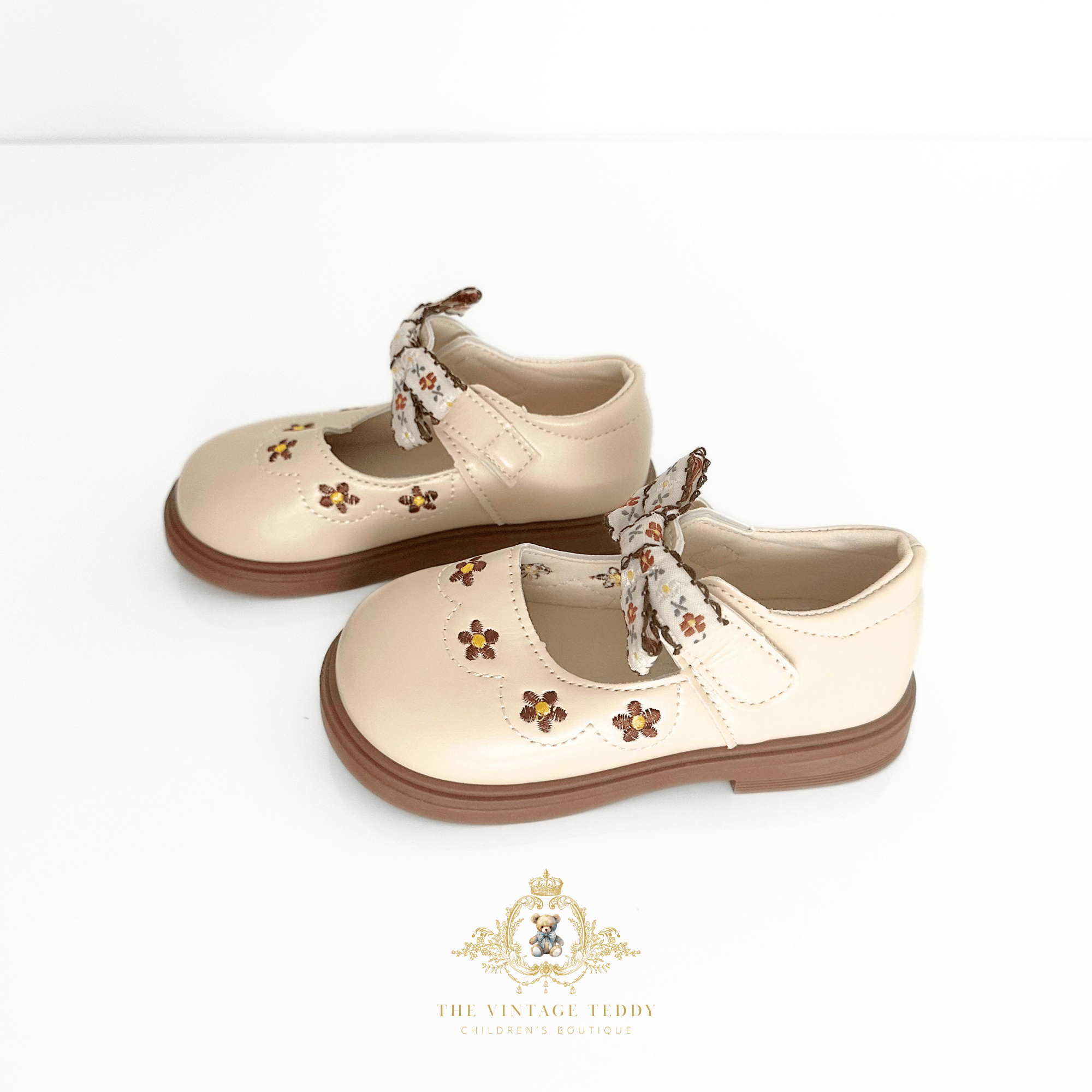 A pair of beige Mary Jane style children's shoes with floral brogue details and ribbon bows, displayed on a clean white background with a brand watermark at the bottom.
