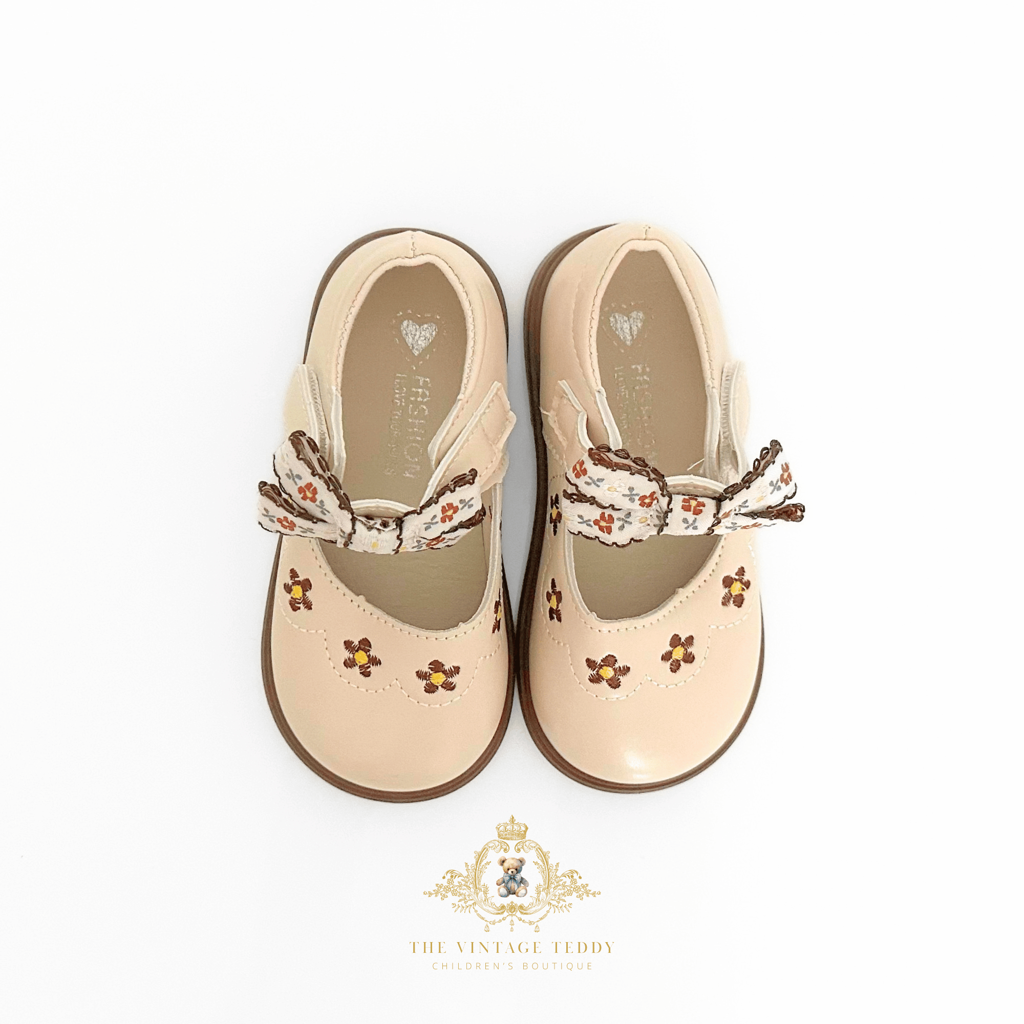 A pair of beige Mary Jane style kids shoes for toddlers, featuring embroidered floral details and a decorative ribbon bow on the strap, shown from a top-down view on a white background.