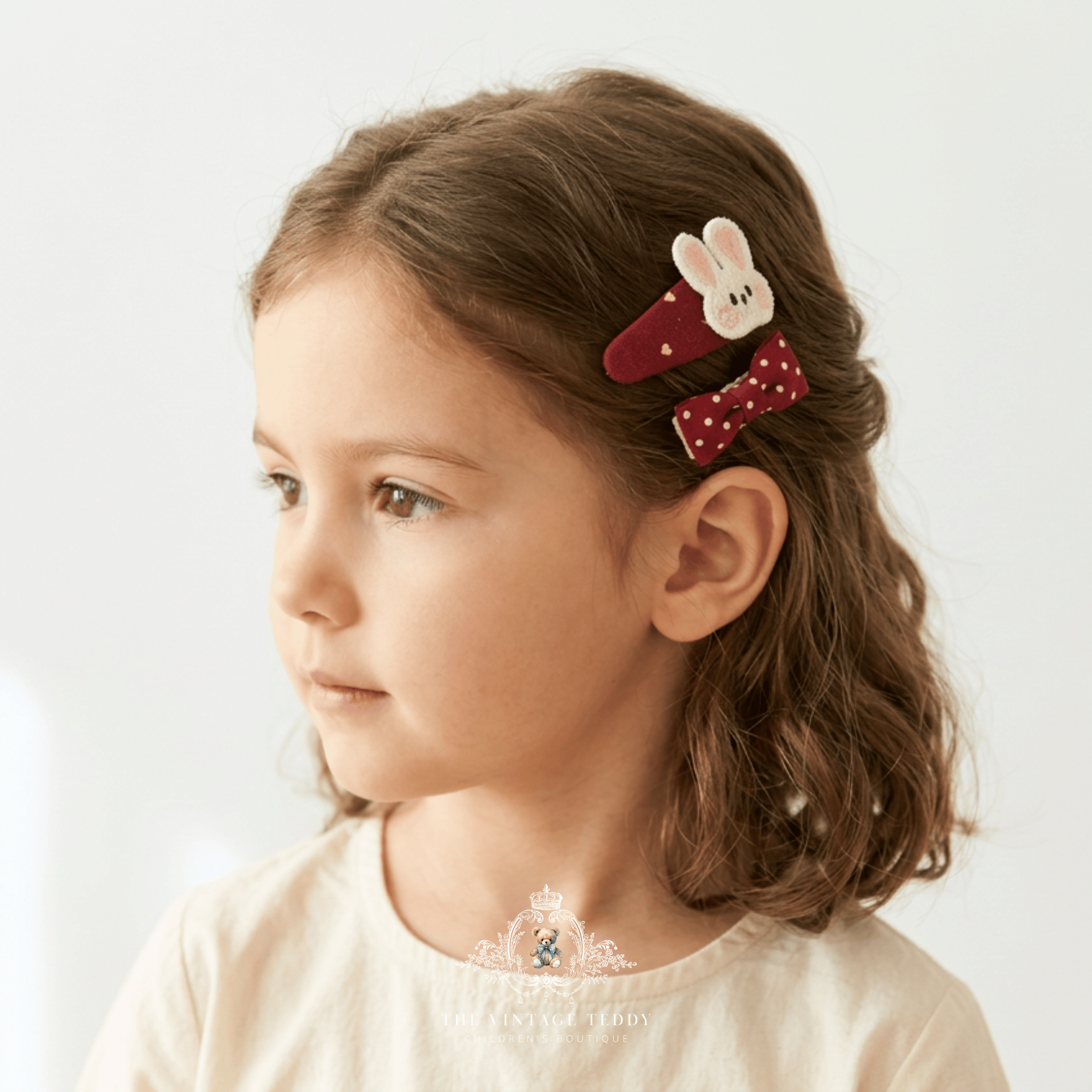 A young girl with brown hair wearing two red and white kids accessory hair clips, one with an embroidered bunny face and the other a polka-dot bow, shown in a close-up profile shot against a light background.