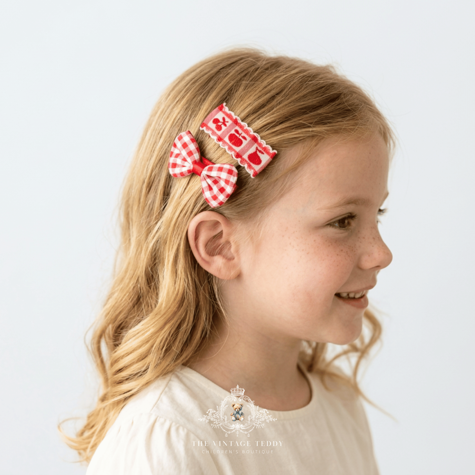 A young girl with blonde hair is shown in profile wearing two red and white gingham bow kids hair clips. The photo is shot against a clean, light gray background with The Vintage Teddy's watermark logo at the bottom.
