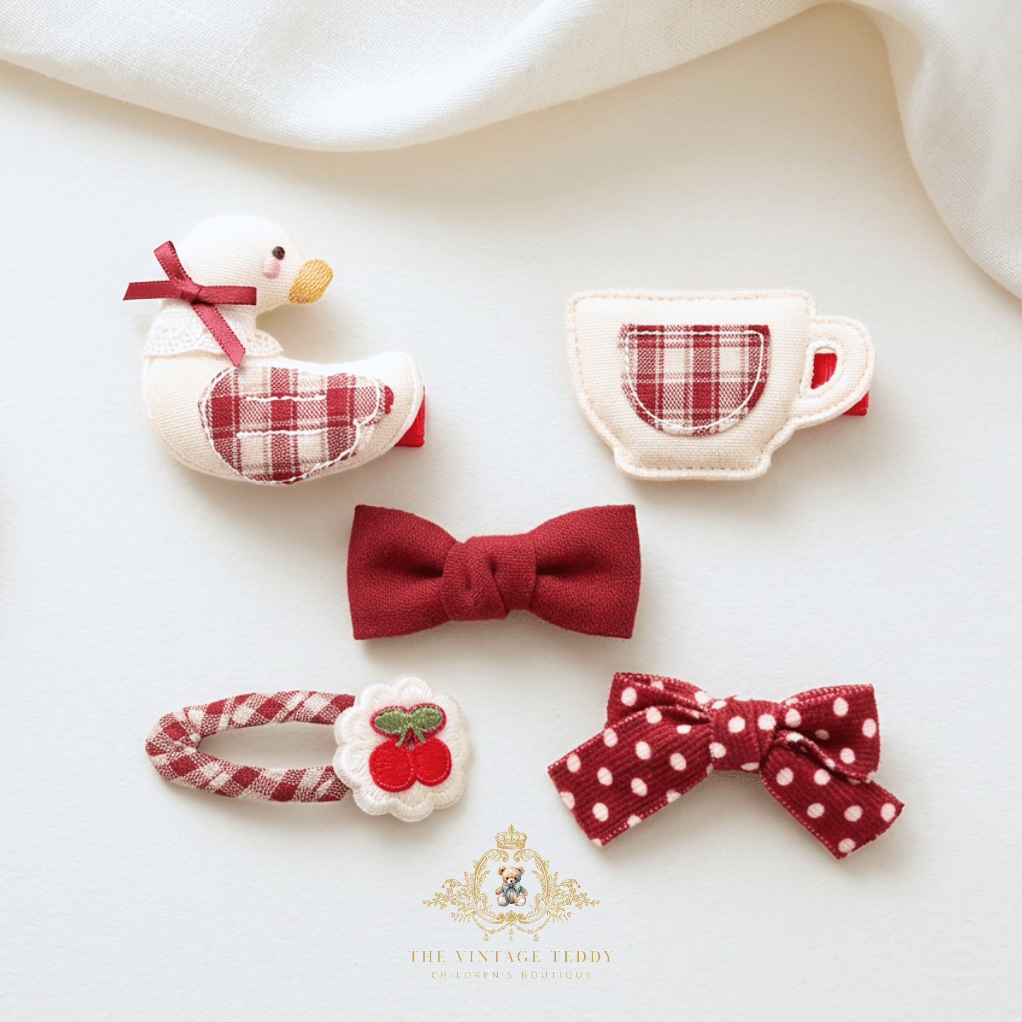 Five handmade plush fabric hair clips accessories for children in a red and white cottage-core theme, including a goose, teacup, bow, polka dot bow, and cherry hair clip-on, arranged on a white fabric background. A logo is visible at the bottom that reads The Vintage Teddy with a cute teddy bear mascot.