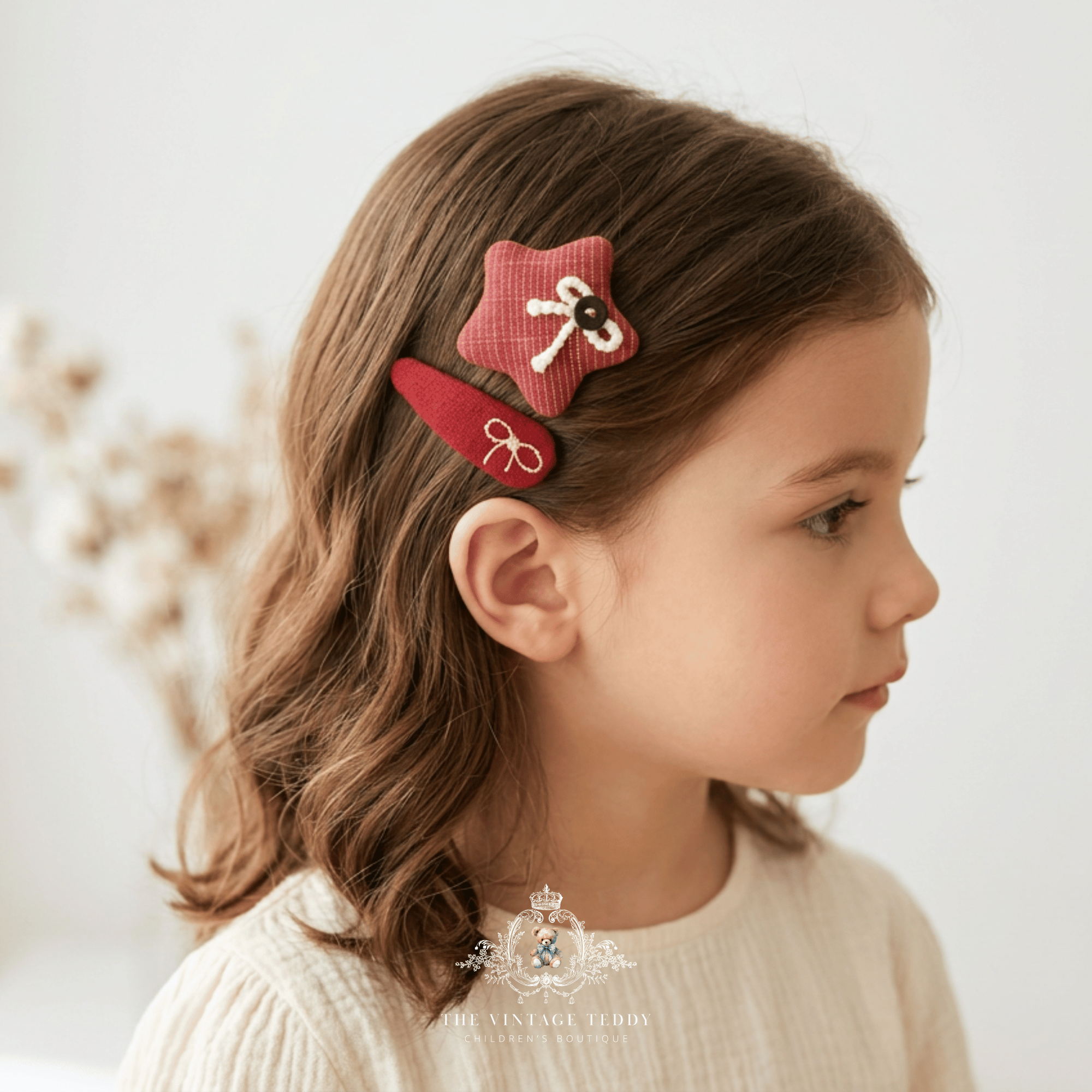 A young girl with brown hair is shown in profile wearing two fabric kids hair clips, one star-shaped and one elongated in red, both decorated with white bow details. The background is a soft, bright, neutral studio setting with blurred dried flowers.