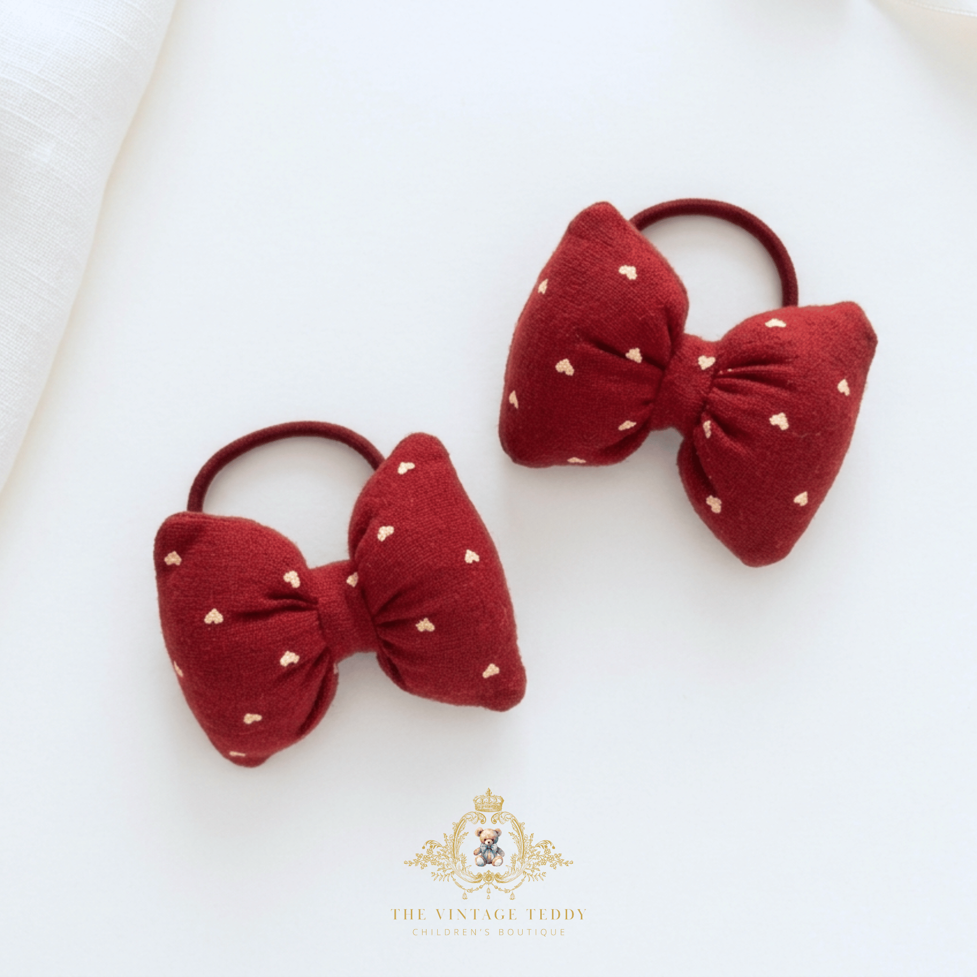 Two red fabric kids bow ponytail hair ties with small white heart patterns, displayed on a clean white background with The Vintage Teddy's  brand watermark at the bottom.