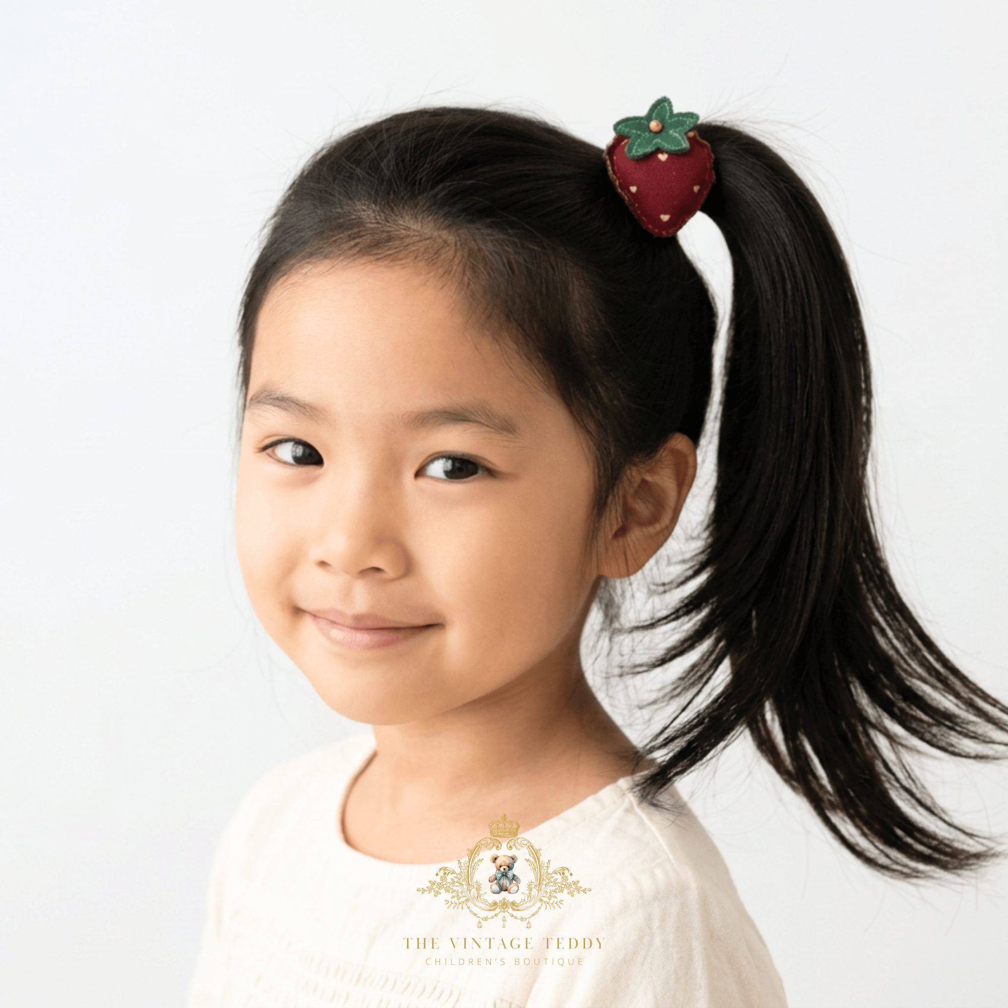 A young girl with a dark ponytail wearing a strawberry-shaped hair tie, photographed against a clean white background. A watermark for 'The Vintage Teddy' is visible at the bottom.