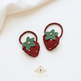 A pair of strawberry-shaped hair ties made from dark red felt with green felt leaves and small gold-colored seed details, displayed on a clean white background with a soft cream fabric corner and a brand watermark at the bottom.