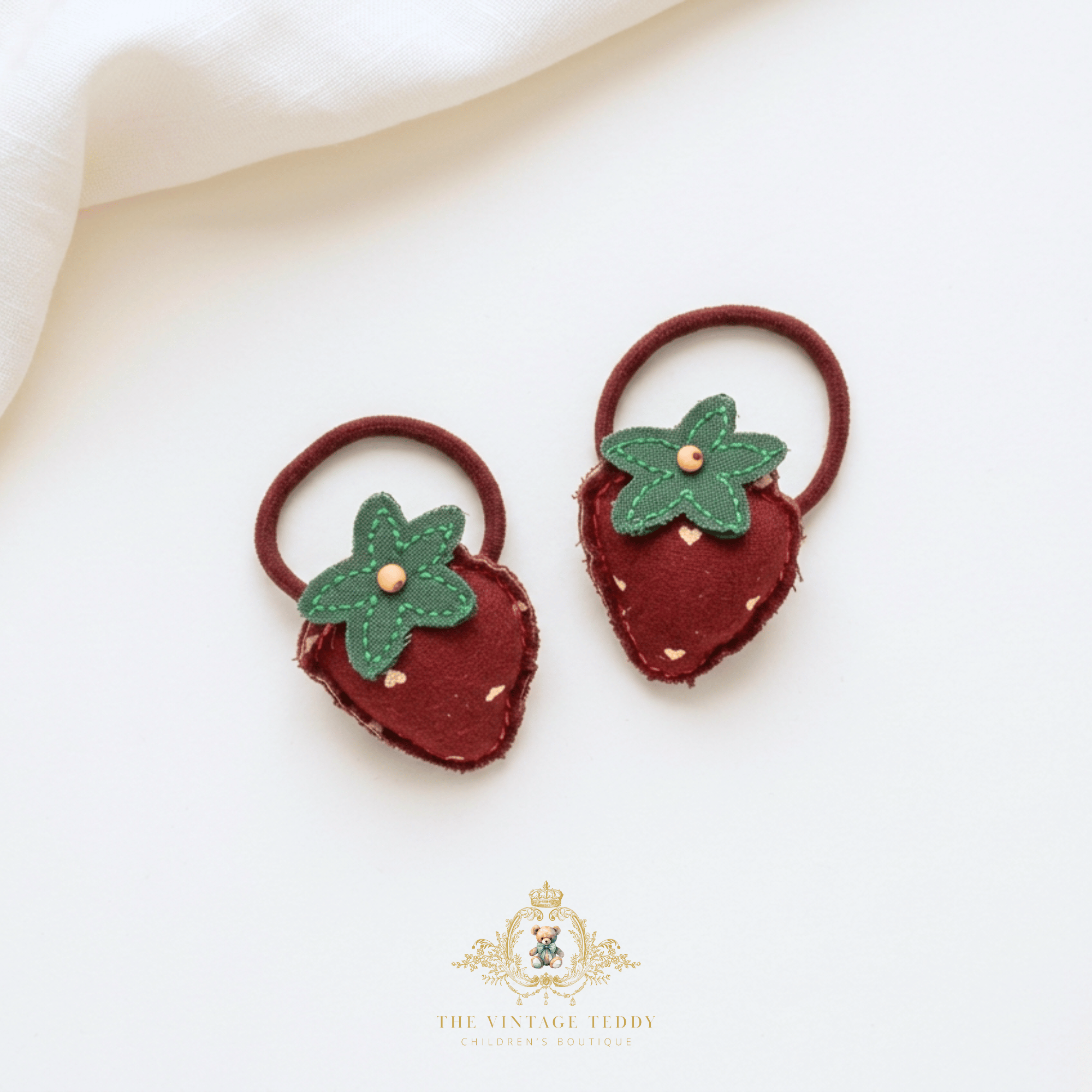 A pair of strawberry-shaped hair ties made from dark red felt with green felt leaves and small gold-colored seed details, displayed on a clean white background with a soft cream fabric corner and a brand watermark at the bottom.