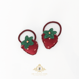 A pair of strawberry-shaped hair ties on a white background, featuring a textured red felt strawberry body with a green felt leafy top and a small gold bead in the center, attached to dark red elastic bands. The brand logo 'The Vintage Teddy' is visible at the bottom.