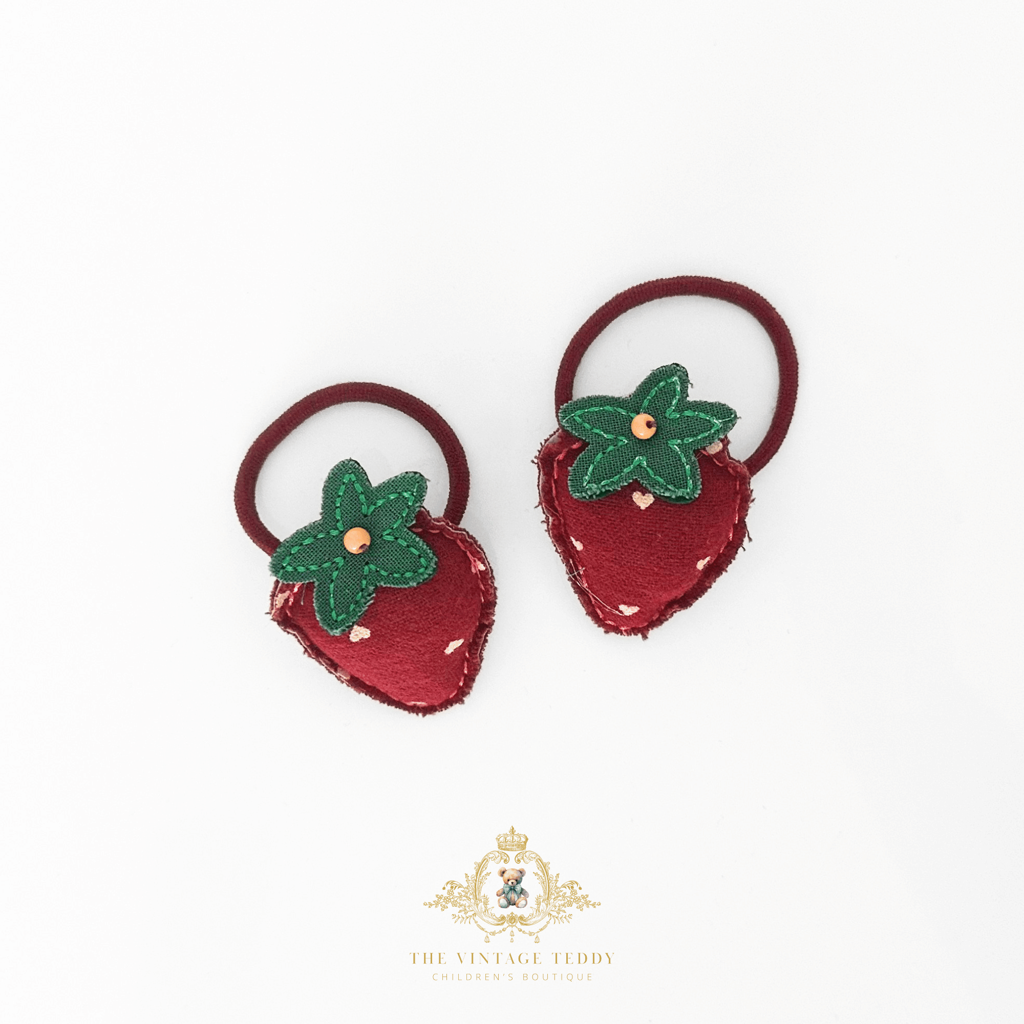A pair of strawberry-shaped hair ties on a white background, featuring a textured red felt strawberry body with a green felt leafy top and a small gold bead in the center, attached to dark red elastic bands. The brand logo 'The Vintage Teddy' is visible at the bottom.