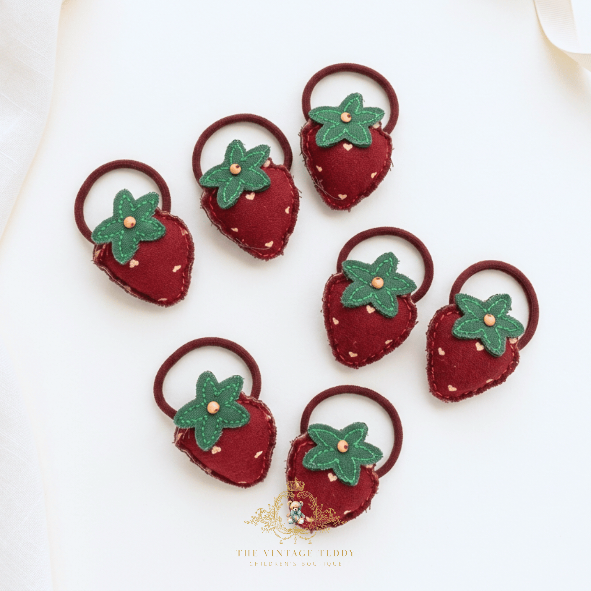 7 strawberry shaped plush little girls hair ties accessories for kids, featuring crimson red fabric with little white hearts, green stem with a gold bead, and elastic band set  on a creamy vanilla surface with The Vintage Teddy's logo bottom-center.