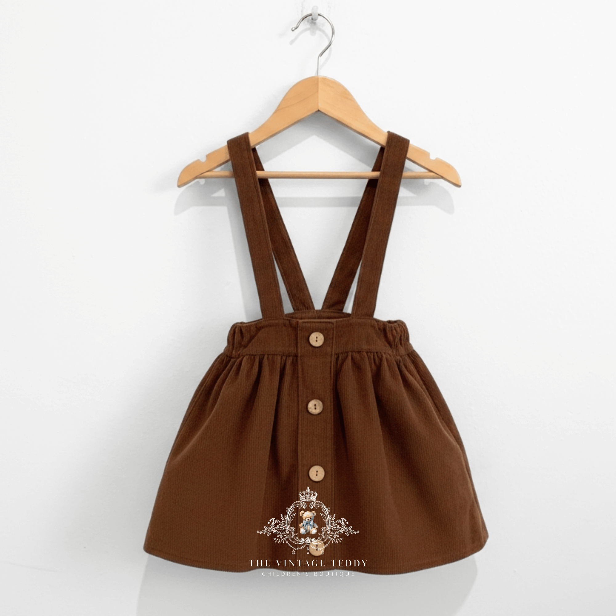 A brown corduroy fabric baby or toddler skirt with adjustable suspenders and with buttons at the front, hanging on a wooden hanger against a plain white wall. The Vintage Teddy watermark in white color at the bottom center.