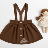 A fall brown girl's corduroy skirt with suspenders, featuring buttons at the front. The corduroy skirt is displayed flat on a white background next to a small handmade doll. The Vintage Teddy logo in white at the bottom.