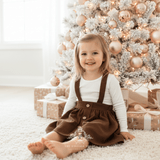 A young child sitting on a white shag rug in front of a decorated Christmas tree, smiling and wearing a corduroy suspender children's skirt in a brown color over a white long-sleeve top, white tights, with wrapped gifts and presents under the tree. A logo is visible at the bottom with teddy bear, crest and The Vintage Teddy text.