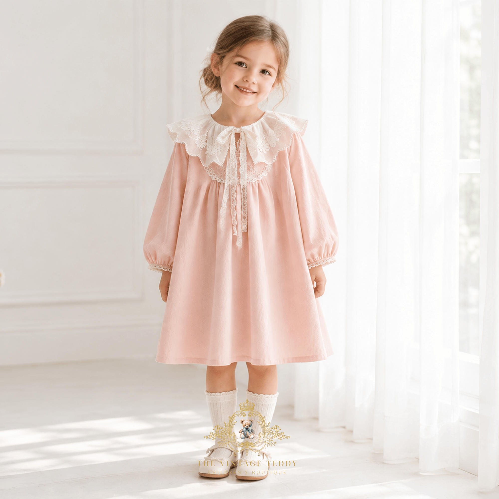 A young girl with hair swept up in a side bun stands smiling in a blush pink children's dress with a lovely vintage-style  lace bib collar paired with white Mary Jane shoes, in light an airy room next to a window with sheer white curtains.