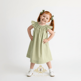 A young girl with red hair in pigtails wearing a sage green smocked dress, standing with hands on hips against a white studio background. The brand logo 'The Vintage Teddy' is visible at the bottom.