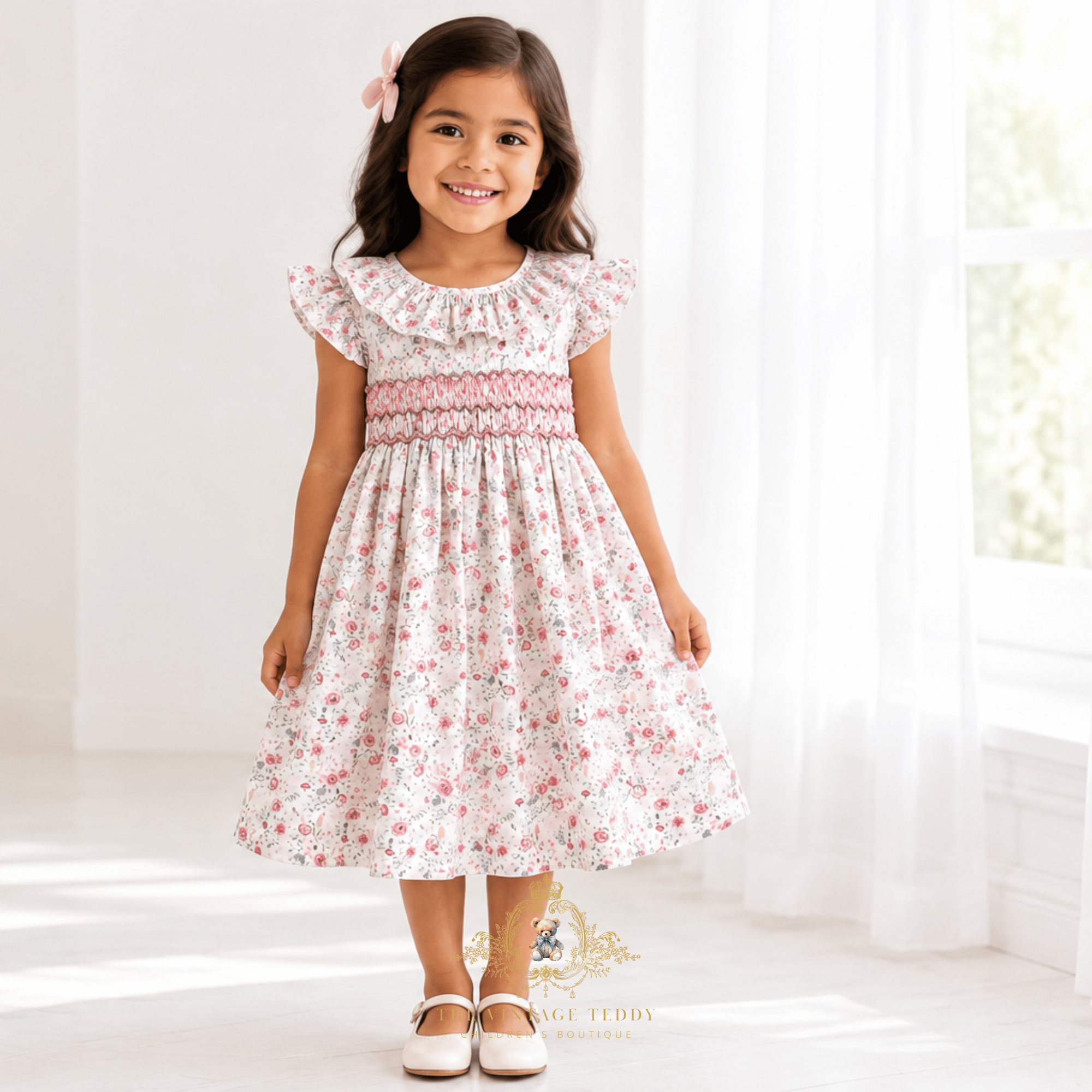 A young girl wearing a white smocked dress with a pink and white floral pattern, standing in a bright, sunlit room with white curtains.