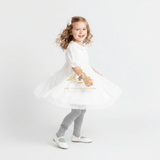 A young girl with curly light brown hair is wearing a white dress, grey knit tights, and white Mary Jane shoes, posing in a twirl on a seamless white studio background. A watermark for 'The Vintage Teddy' is visible on the dress.