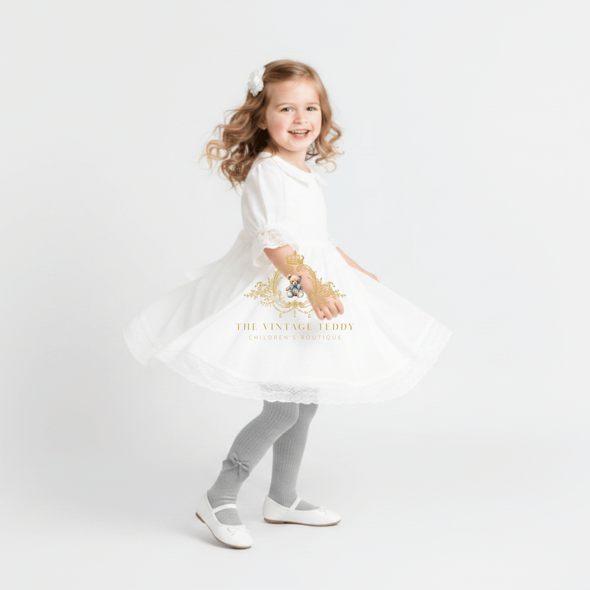 A young girl with curly light brown hair is wearing a white dress, grey knit tights, and white Mary Jane shoes, posing in a twirl on a seamless white studio background. A watermark for 'The Vintage Teddy' is visible on the dress.