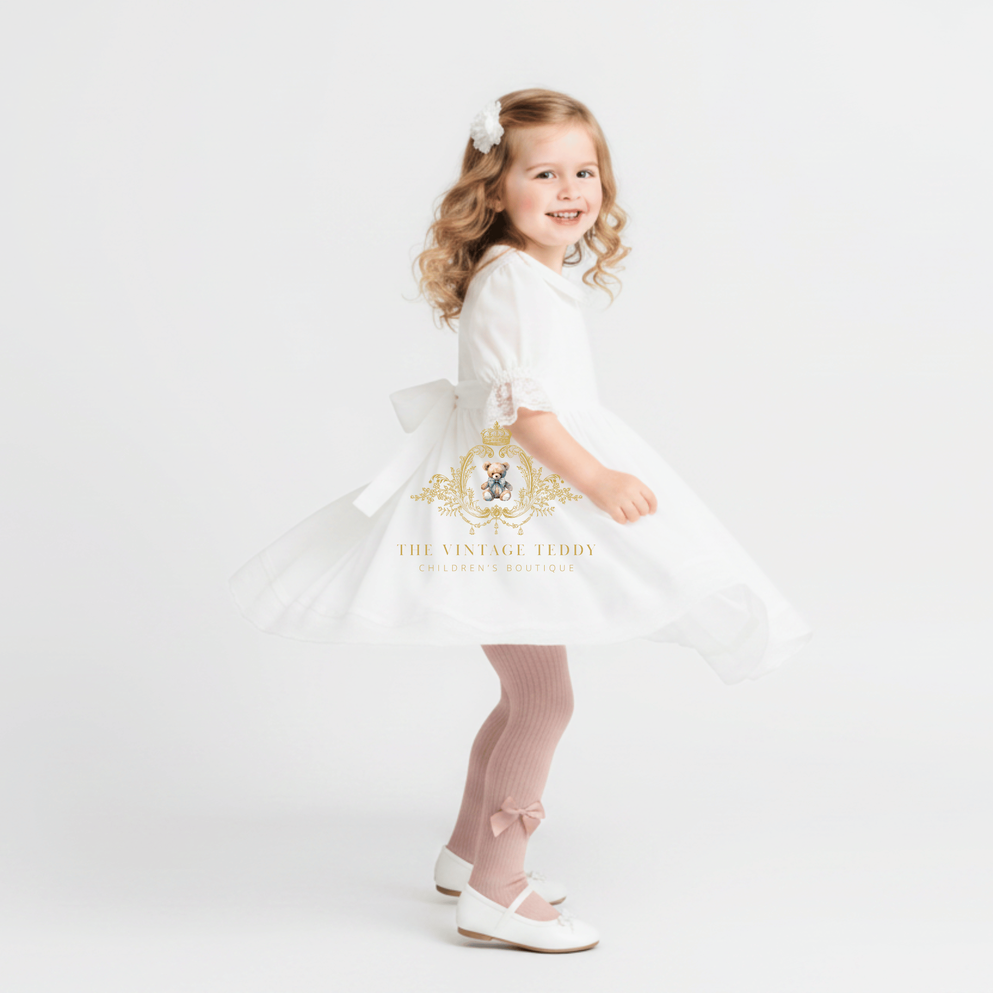 A young girl with blonde curly hair wearing a white dress, dusty pink tights, and white shoes, posing in a studio setting. The dress features a white collar, short sleeves, and a flared skirt with a large bow at the back.