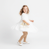 A young girl with curly light brown hair is wearing a white dress, cream-colored knit tights, and white Mary Jane shoes, posing in a twirl on a seamless white studio background. A watermark for 'The Vintage Teddy' is visible on the dress.