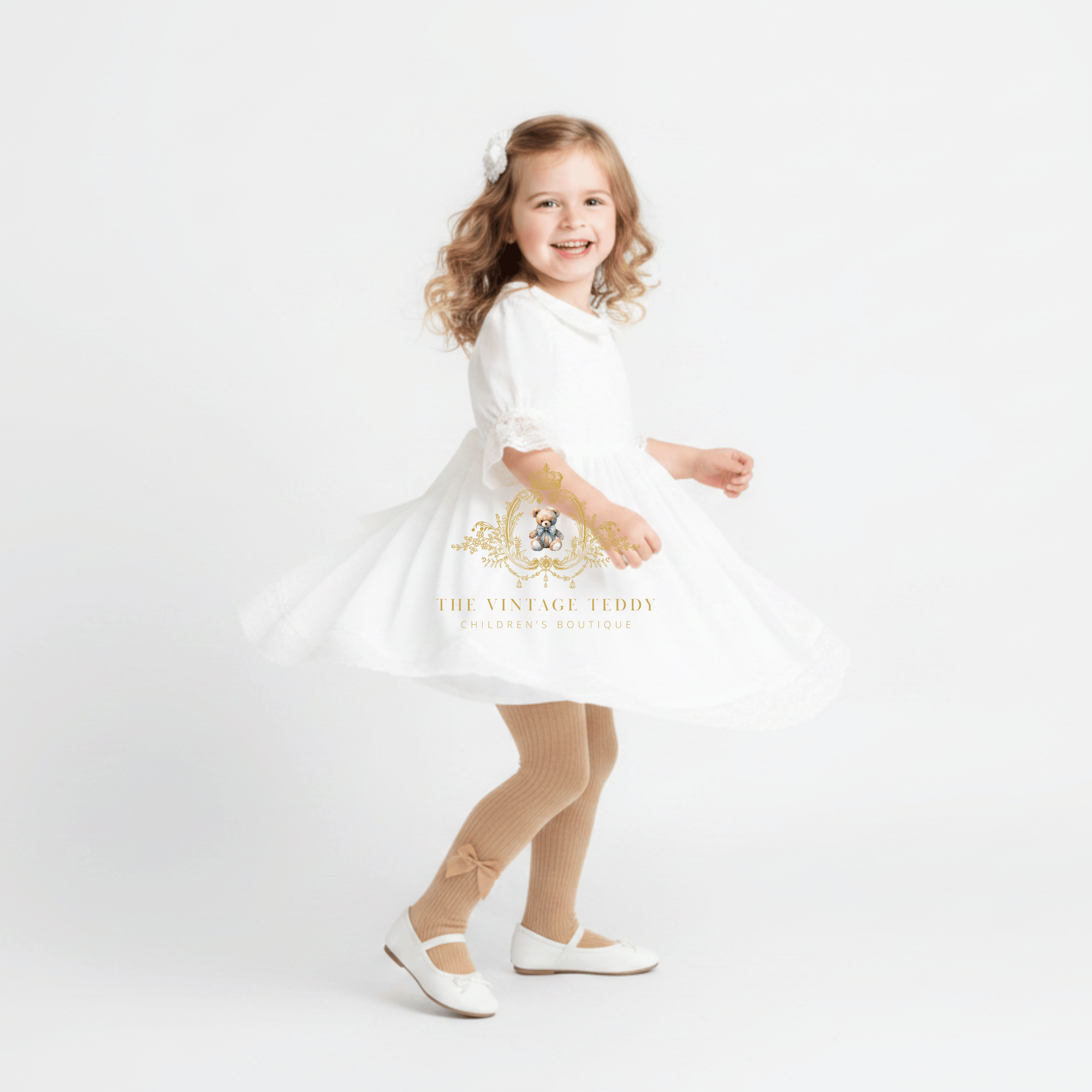 A young girl with curly light brown hair is wearing a white dress, cream-colored knit tights, and white Mary Jane shoes, posing in a twirl on a seamless white studio background. A watermark for 'The Vintage Teddy' is visible on the dress.