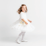 A young girl with curly light brown hair wearing a white dress, white tights, and white shoes, twirling in a white studio setting. The dress features a gold embroidered crest and the text 'THE VINTAGE TEDDY' on the front.