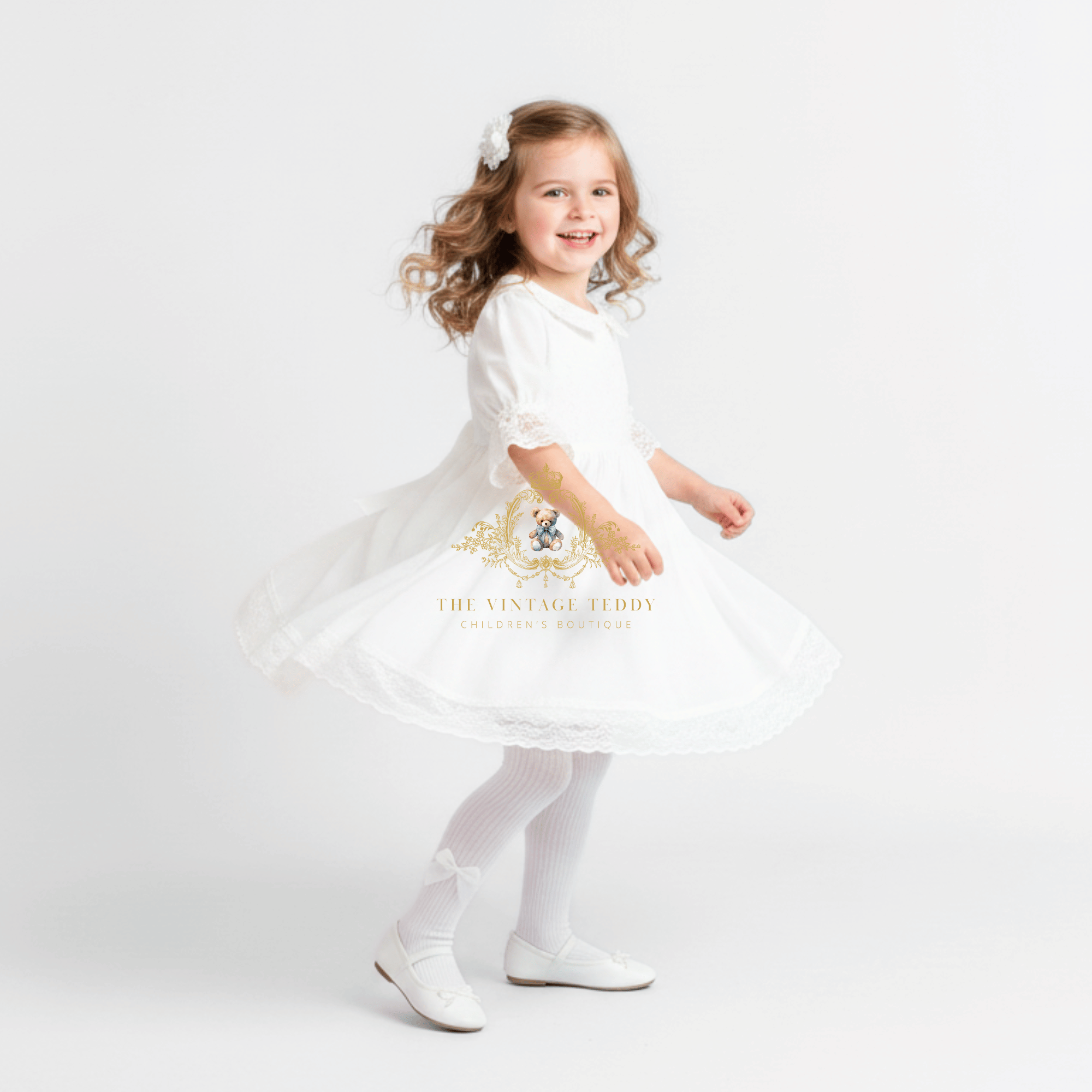 A young girl with curly light brown hair wearing a white dress, white tights, and white shoes, twirling in a white studio setting. The dress features a gold embroidered crest and the text 'THE VINTAGE TEDDY' on the front.