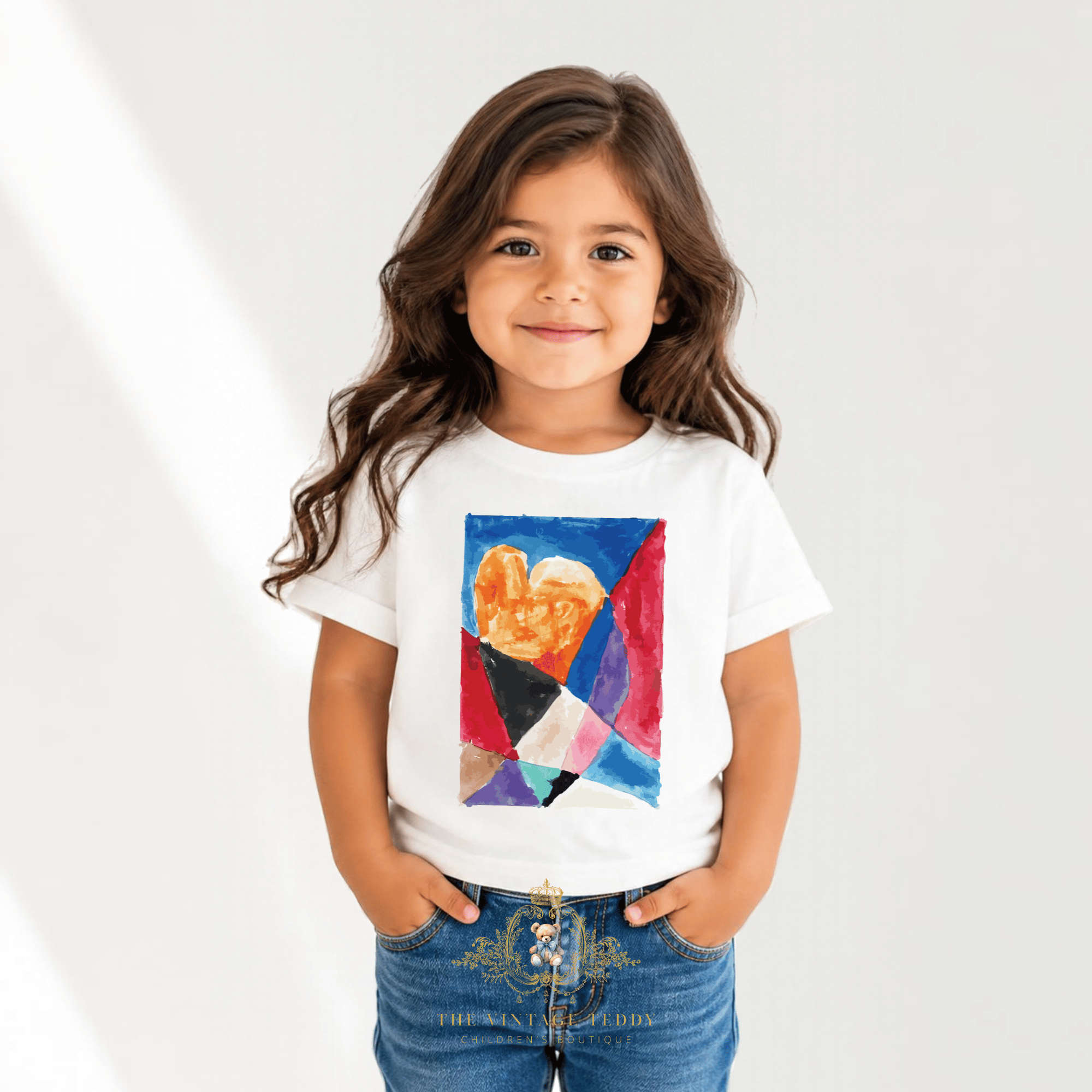 A smiling little girl with long wavy brown hair wearing a white kids t-shirt with a custom designed colorful stain glass watercolor with an abstract heart in the center and blue jeans, on a airy light studio background.